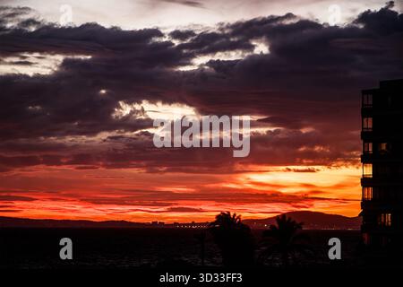 Dramatic coastal sunset with vivid red and orange clouds over the sea and silhouetted building and palm trees can pastilla mallorca balaeric isles Stock Photo