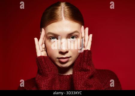 Red-haired young woman in a stylish winter sweater poses gracefully on a striking red background. Stock Photo