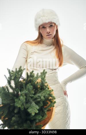 Redheaded woman wearing a fur hat stands against a white background holding a Christmas tree. Stock Photo