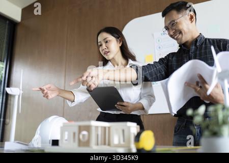 Engineers enthusiastically discussing sustainable architecture plans while reviewing models and blueprints in a modern office Stock Photo