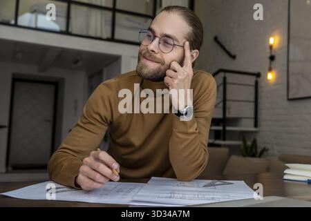 Portrait of pensive bearded young engineer architect in glasses sitting at the table and creating blueprint thinking about new project Stock Photo