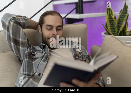Cheerful relaxed man reading book while sitting on cough at home, enjoying interesting novel Stock Photo