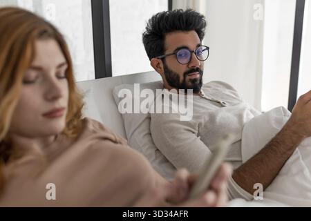 Young Mixed-race couple with serious expression using smartphone lying on bed Stock Photo