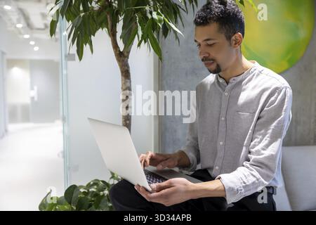 Concentrated young African American company employee seated on the sofa typing on the portable computer Stock Photo