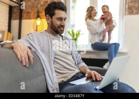 Father working on laptop, mother and daughter on background playing in home interior Stock Photo
