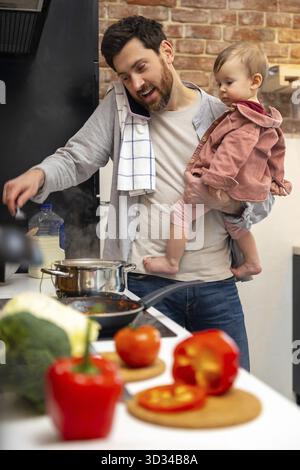 Multitasking father babysitting, cooking breakfast and talking on smart phone at home in kitchenn Stock Photo
