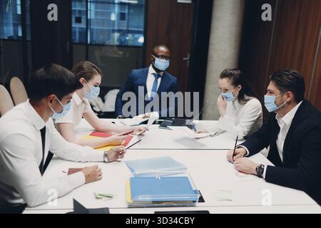 Business team in medical face mask at office meeting. Business people group conference discussion with boss Stock Photo