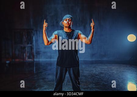 Young man break dancer standing in dark hall on wall background. Tattoo on hands Stock Photo