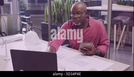 Reviewing senior architect wearing glasses and red shirt at office desk, with laptop and blueprints Stock Photo