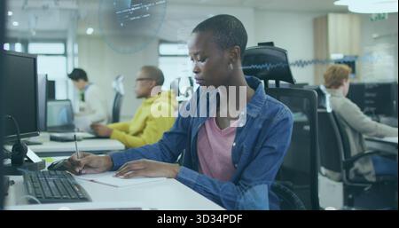 Woman writing on notepad in open office with coworkers coding on computers, holographic overlays Stock Photo