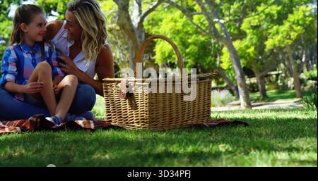Sitting mother and daughter talking on plaid blanket in park with wicker picnic basket, copy space Stock Photo