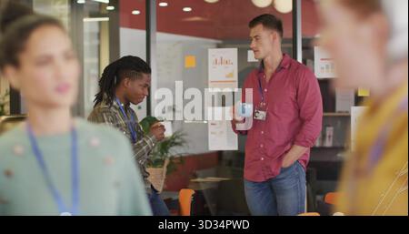 Chatting two male co-workers holding coffee mugs in office lounge, with glass wall charts and plant Stock Photo