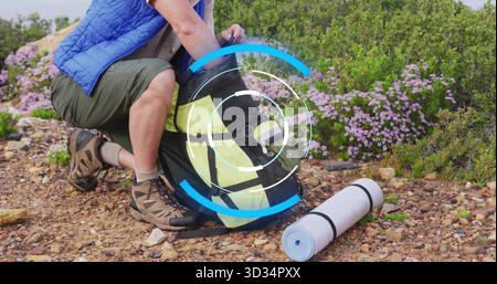 Unpacking male hiker wearing blue vest organizing backpack on hillside, with rolled sleeping mat Stock Photo