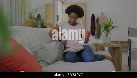 Reading woman holding pink mug, turning book pages on light sofa in living room, with plants Stock Photo