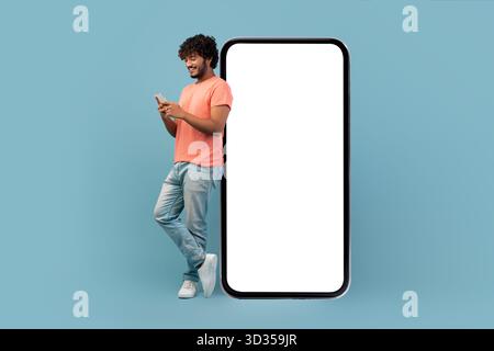 Cool Indian Guy Leans on a Giant Smartphone While Texting and Smiling in Blue Studio Setting Stock Photo