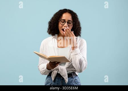 Black Female Student Yawns While Studying for University Exam in Blue Studio Stock Photo