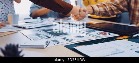 A close-up of a handshake between two professionals in a bright office, symbolizing a successful partnership. Project documents and charts lay Stock Photo