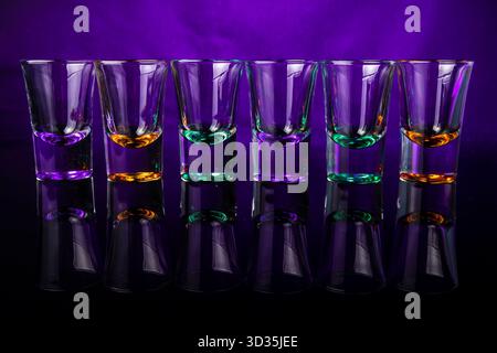 Six empty shot glasses with colored bases arranged in a line on a purple background. Reflections and symmetry create a vibrant composition. Stock Photo