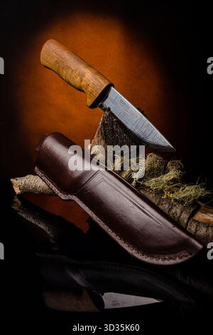 Artisan knife with polished steel blade and wooden handle, standing in a log. Leather sheath below on reflective surface. Stock Photo
