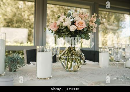 A beautifully arranged table showcases a glass vase filled with pink and white roses, surrounded by tall glasses and a lace tablecloth, all illuminate Stock Photo