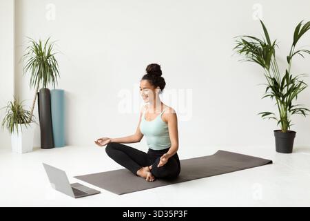 Meditating Young Woman Practicing Remote Yoga in a Calm Indoor Space Near a Laptop Stock Photo