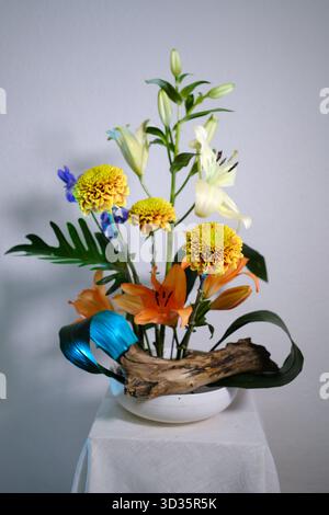 Colorful arrangement of flowers in a white vase, featuring lilies, marigolds, and blue accents, displayed on a soft gray background that highlights th Stock Photo
