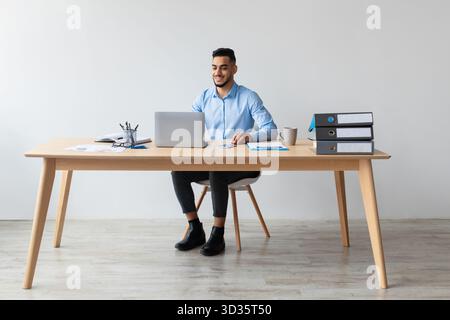 Middle Eastern Man Working on Laptop at Modern Office Desk With Cheerful Demeanor Stock Photo
