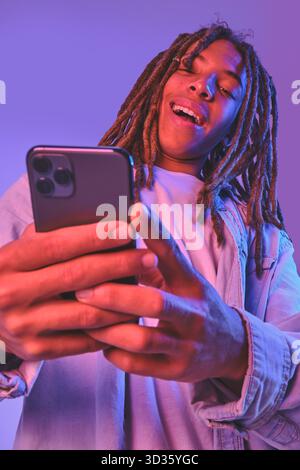 Young man with dreadlocks smiling while holding and using a smartphone Stock Photo