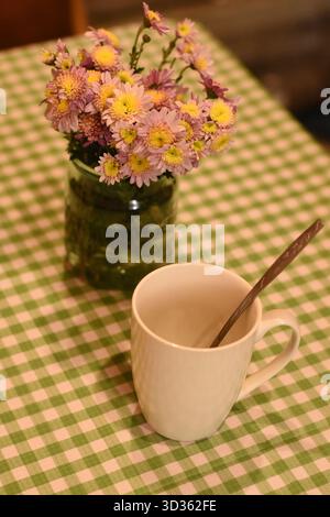 White cup with a spoon on a green checkered tablecloth, next to a glass jar with pink chrysanthemums. Top view, cozy home-style photo. Stock Photo