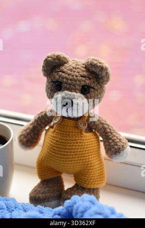 Brown knitted teddy bear on windowsill with blue blanket, cup of coffee and red blurred background Stock Photo