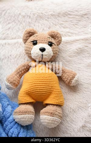 Brown knitted teddy bear in yellow jumpsuit lying on gray blanket with part of blue knitted blanket visible Stock Photo