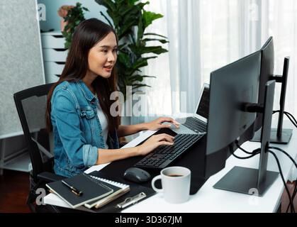 Smiling young Asian IT developer working software development information on pc screens and laptop, creating program coding for latest version Stock Photo