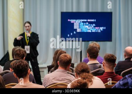 Speaker presents to seated audience at tech conference while displaying code on large screen. Attendees look attentive, notes ready, room bright with soft curtains and modern equipment  Stock Photo