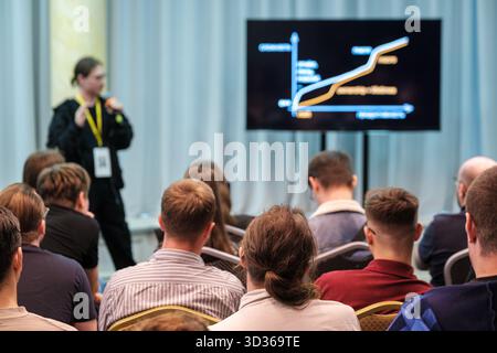 Speaker presents data to seated audience in conference room with large screen showing chart Stock Photo