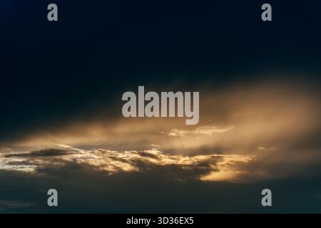 Dramatic clouds illuminated by golden sunlight stretch across the sky, creating a peaceful and atmospheric sunset view above the horizon. Stock Photo