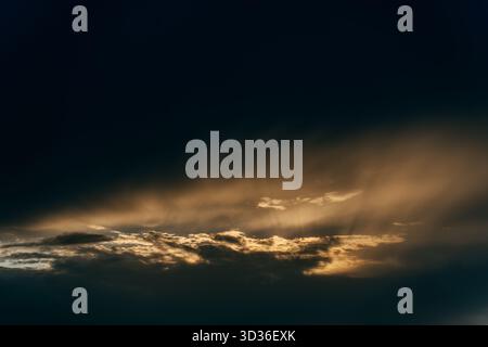 Soft beams of light filter through thick clouds as the sun sets, casting warm highlights across a vast, peaceful sky. Stock Photo