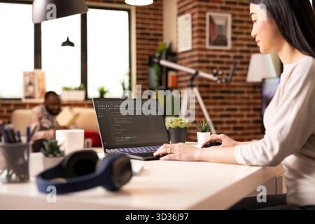 Asian female programmer sits at home desk, writing and debugging coding scripts on laptop screen. Woman entrepreneur working remotely on app development for startup tech company. Stock Photo