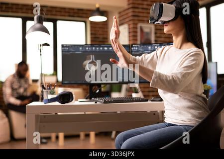Female engineer wearing VR headset and gesturing hands in 3D simulation to review clamp model. Young woman works remotely with advanced technology, doing interactive engineering inspection on tools. Stock Photo