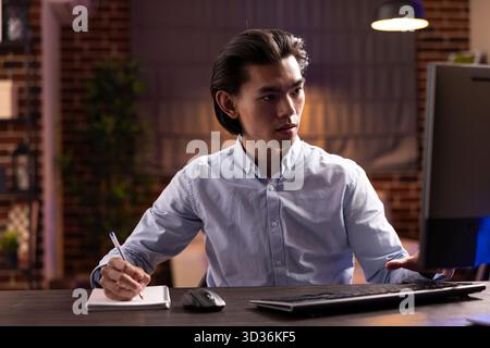 Serious and focused businessman looks at computer screen while writing notes. Working remotely or studying online, asian entrepreneur conducts research or prepares for exam using desktop PC. Stock Photo