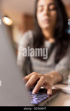 Focused woman working on laptop, writing business blog and analyzing financial insights from home. Close up of indian female professional gathering data on digital device at desk. Stock Photo