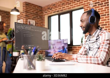 Black male programmer smiling at desktop monitor and reviewing software code. Confident developer working remotely on secure website for startup tech company from home office. Stock Photo