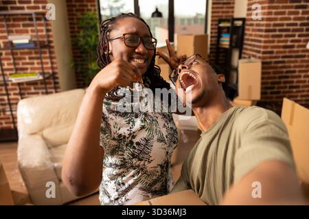 Cheerful boyfriend and girlfriend take playful selfie together, holding keys and cardboard box after moving into new home. Man holds phone, making goofy face and enjoying moment with partner. Stock Photo