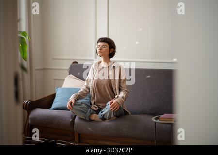 Young woman meditating on the sofa, calming mind and practicing inner peace, caring for mental health stability. Stock Photo