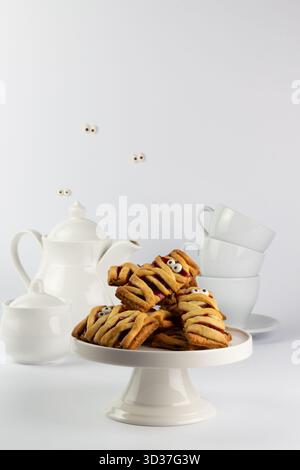 Mummy-themed cookies with cute eyes are neatly arranged on a white cake stand. A white tea set sits in the background, creating a fun Halloween scene. Stock Photo