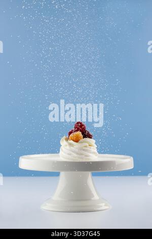 A delicious dessert features whipped cream topped with fresh fruit, placed on a white cake stand. Sugar is being sprinkled over the treat against a bl Stock Photo