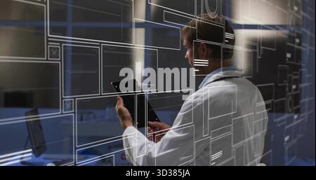Tech wearing lab coat holding tablet monitoring overlay in control room, with laptop and monitors Stock Photo