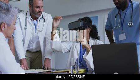 Lab coat doctor wearing VR headset gesturing in hospital conference room, with laptop, stethoscopes Stock Photo
