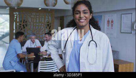 Smiling woman doctor wearing stethoscope in hospital lounge, colleagues using laptop and papers Stock Photo
