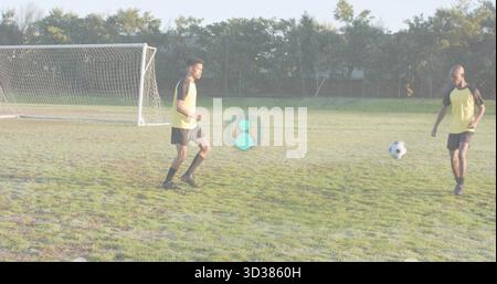 Practicing two male soccer players wearing yellow bibs running on grass pitch beside goal net Stock Photo