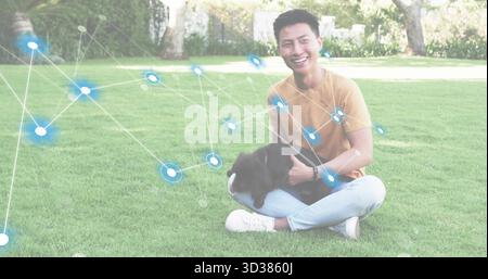 Smiling adult man wearing mustard t-shirt and jeans sitting on park lawn, with black dog Stock Photo